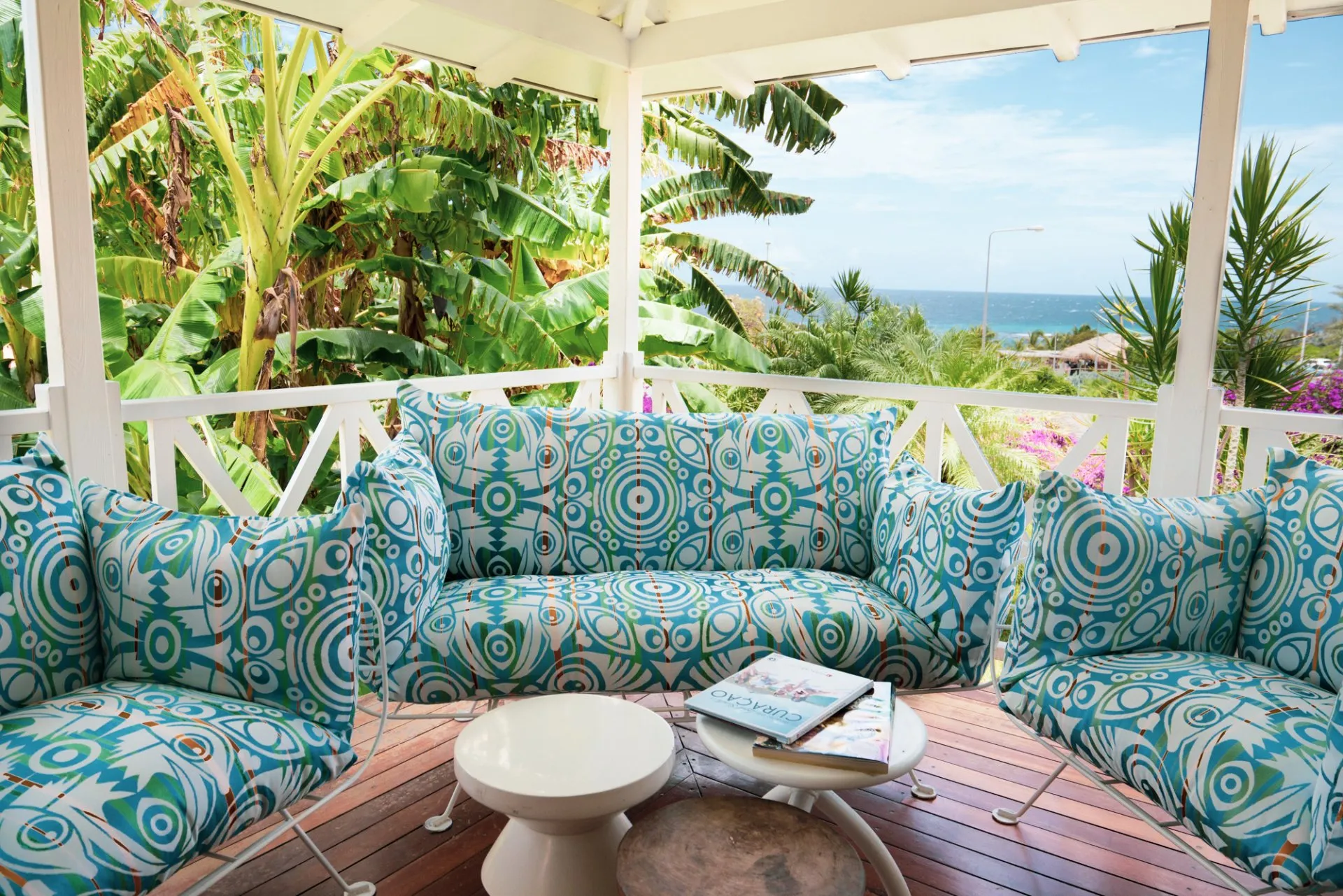 Cozy terrace of a Papagayo Curaçao resort villa featuring vibrant turquoise patterned furniture, surrounded by lush tropical plants and banana trees, with a partial ocean view in the background.