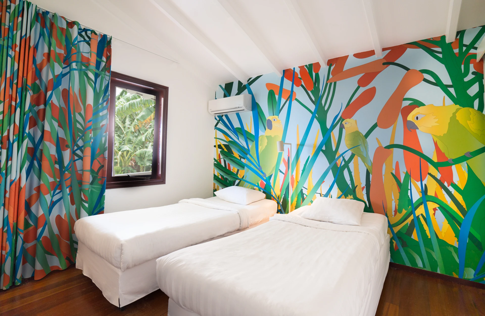 Bright twin-bed villa room at Papagayo Resort featuring colorful tropical parrot-themed wall art, matching vibrant curtains, wooden floors, and a garden view.