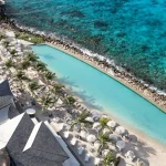 Aerial view of the beachfront infinity pool at Papagayo Beach Hotel in Curaçao, with palm trees, lounge chairs, and turquoise Caribbean Sea in the background.