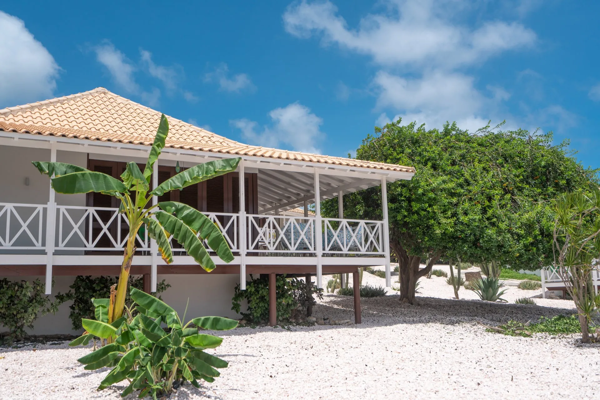 Elevated resort villa at Papagayo Curaçao with a terracotta tiled roof, white wooden railings, surrounding cacti and lush greenery under a partly cloudy sky.