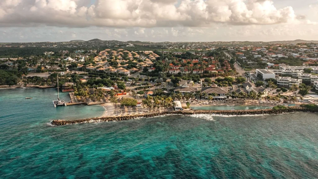 Scenic aerial view of the Jan Thiel area in Curaçao, showcasing a coastal resort with a beachfront infinity pool, surrounding lush villas with thatched roofs, modern white buildings, and the vibrant turquoise Caribbean Sea in the foreground.