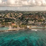 Scenic aerial view of the Jan Thiel area in Curaçao, showcasing a coastal resort with a beachfront infinity pool, surrounding lush villas with thatched roofs, modern white buildings, and the vibrant turquoise Caribbean Sea in the foreground.