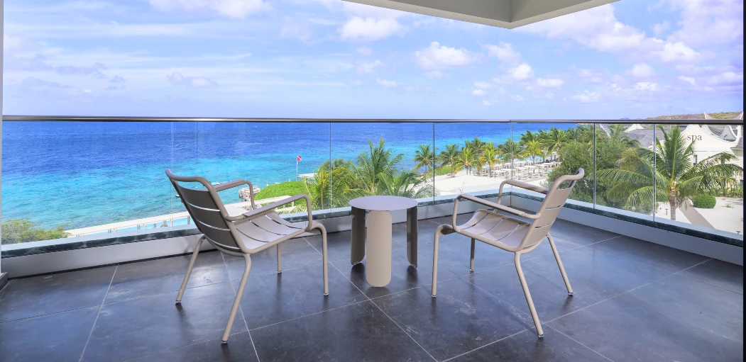 Private balcony of a Papagayo Beach Hotel room featuring modern lounge chairs and a stunning panoramic view of the turquoise Caribbean Sea, palm trees, and the beach below.