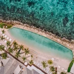Aerial view of the Papagayo Beach Hotel's infinity pool in Curaçao, bordered by palm trees, sun loungers, white sand, and turquoise ocean waters with a rocky shoreline.
