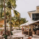 Tropical beachfront lounge area at Papagayo Beach Club in Curaçao, featuring soft bean bag seating on the sand, palm trees, a shaded bar, and white parasols under a sunny sky.