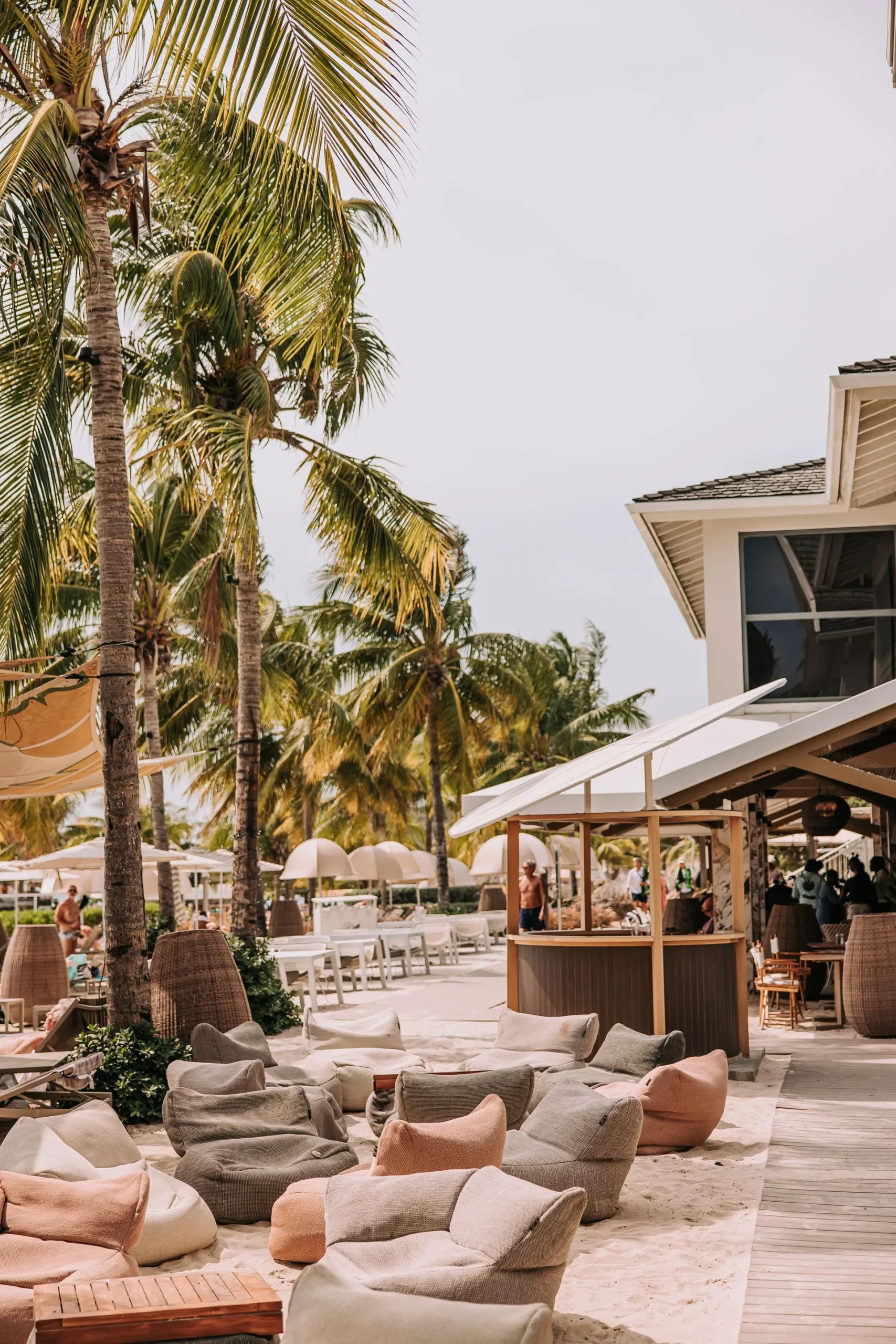 Papagayo Beach Club Tropical beachfront lounge area at Papagayo Beach Club in Curaçao, featuring soft bean bag seating on the sand, palm trees, a shaded bar, and white parasols under a sunny sky.