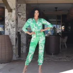 Miss Curaçao 2023 posing confidently at the entrance of Papagayo Beach Club, wearing a vibrant green patterned jumpsuit and high heels, with hands on hips and a radiant smile.