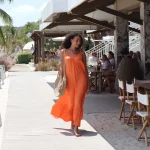 Ona Moody, Miss Netherlands 2022, walks barefoot along the boardwalk at Papagayo Beach Club wearing a flowing orange dress and carrying a straw bag, with restaurant guests seated in the background.