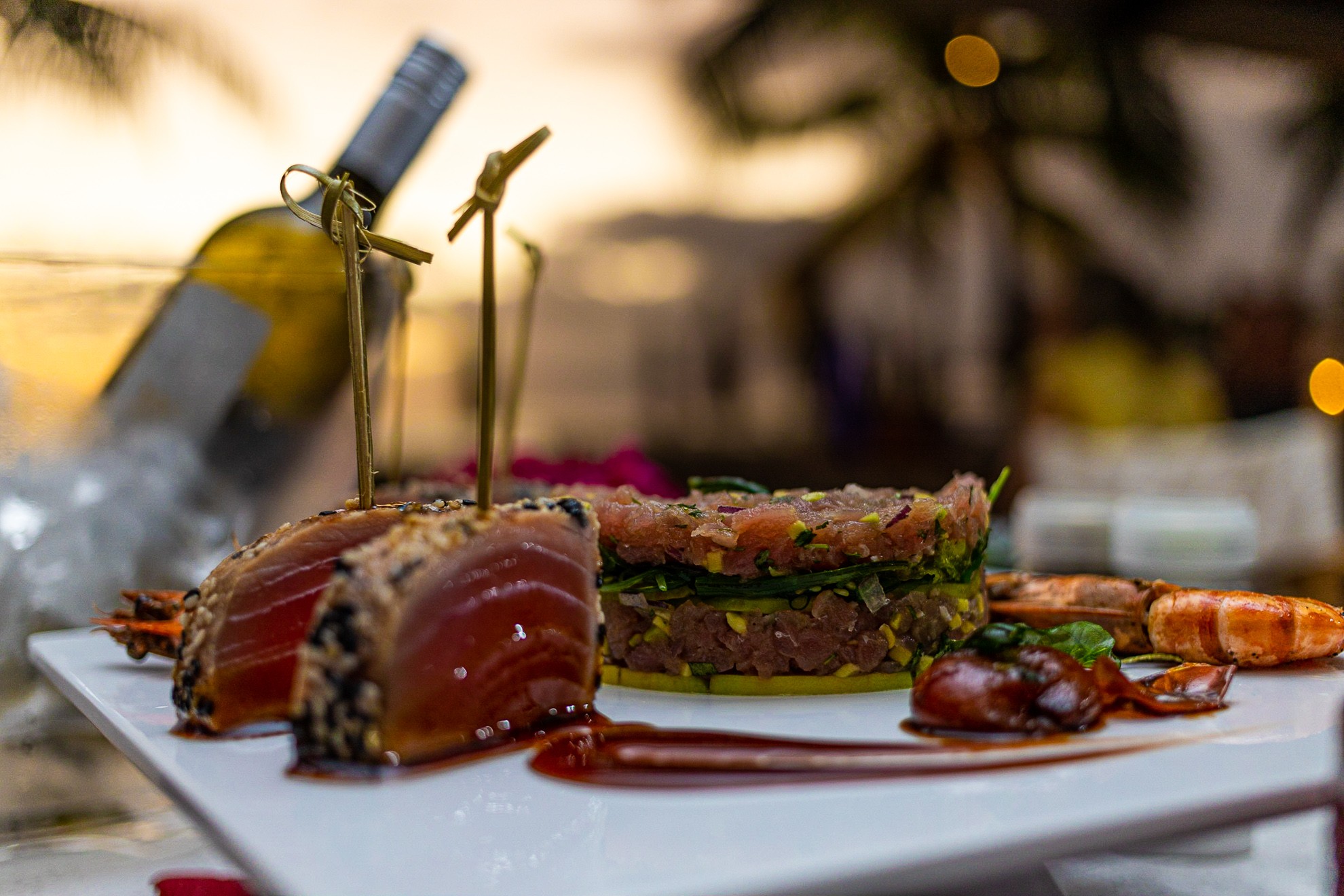 Papagayo Beach Club Restaurant Close-up of a gourmet seafood dish with seared tuna, shrimp, and tartare, served on a white plate with a bottle of white wine in the background at Papagayo Beach Club.
