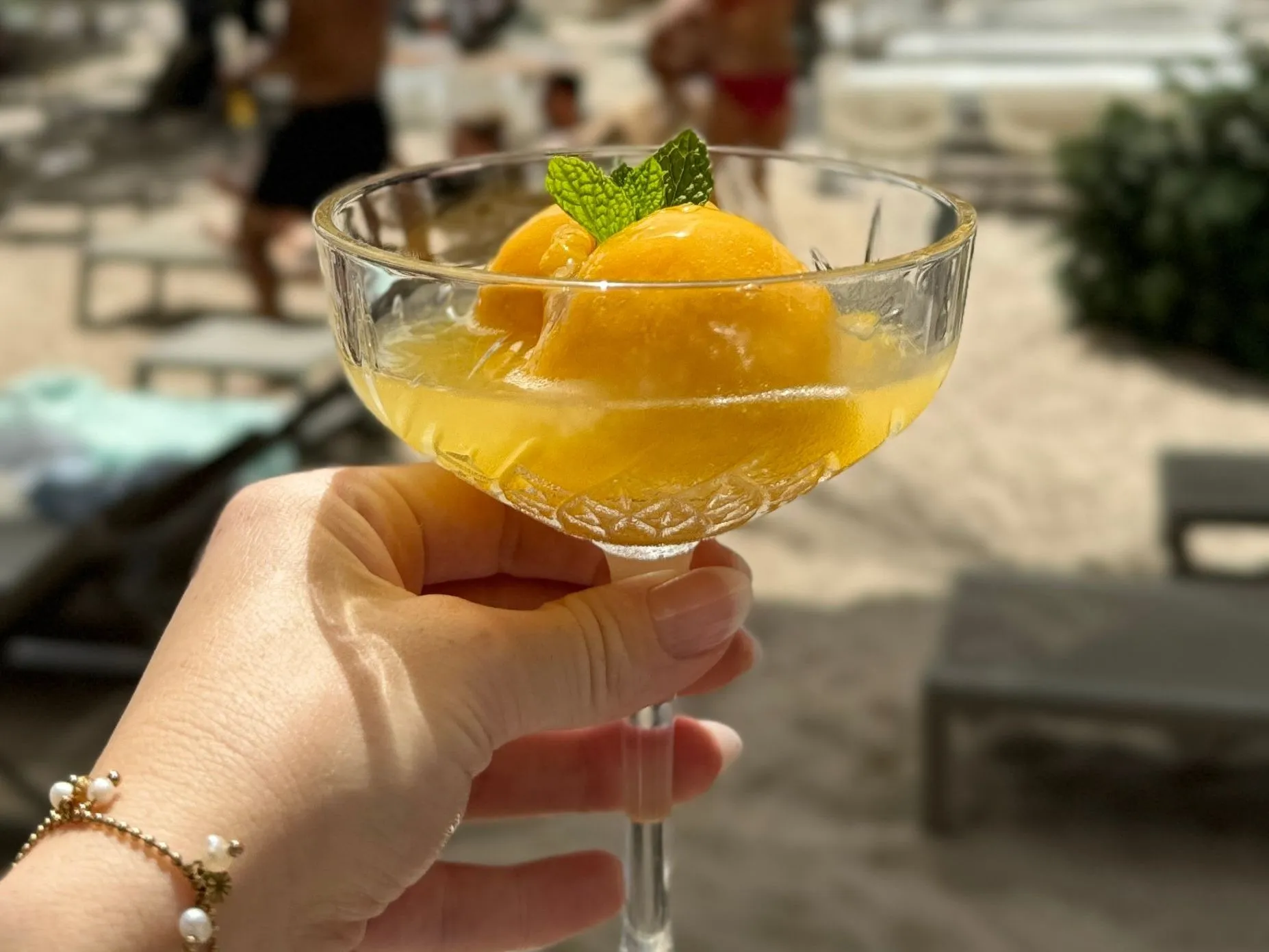 Hand holding a cocktail glass with two scoops of tropical sorbet and a mint leaf garnish at Papagayo Beach Club, with beach chairs and people in the background.