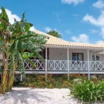 Tropical resort villa at Papagayo Curaçao featuring a wraparound porch, terracotta roof, and lush gardens with banana trees and native plants under a sunny blue sky.