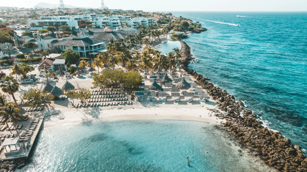Aerial view of Papagayo Beach Club & Hotel in Curaçao, featuring white sand beaches, sun loungers, palm trees, and turquoise waters along the Caribbean coast.