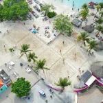 Aerial view of beach tennis courts at Papagayo Beach Curaçao, surrounded by palm trees, sunbeds, colorful beach umbrellas, and nearby turquoise waters where people swim and relax.