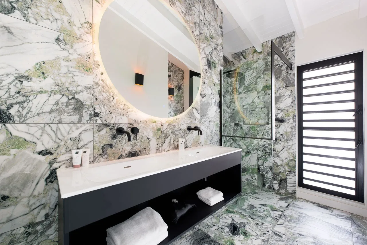 Luxurious bathroom in the Royal Suite Villa at Papagayo Resort, featuring elegant marble walls and floors, a large backlit round mirror, black modern fixtures, and a spacious double sink vanity.
