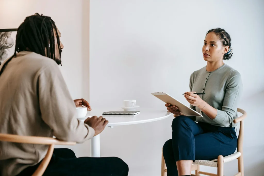 Woman holding a clipboard attentively listening to a man during a one-on-one meeting at a small round table with coffee cups and a notebook