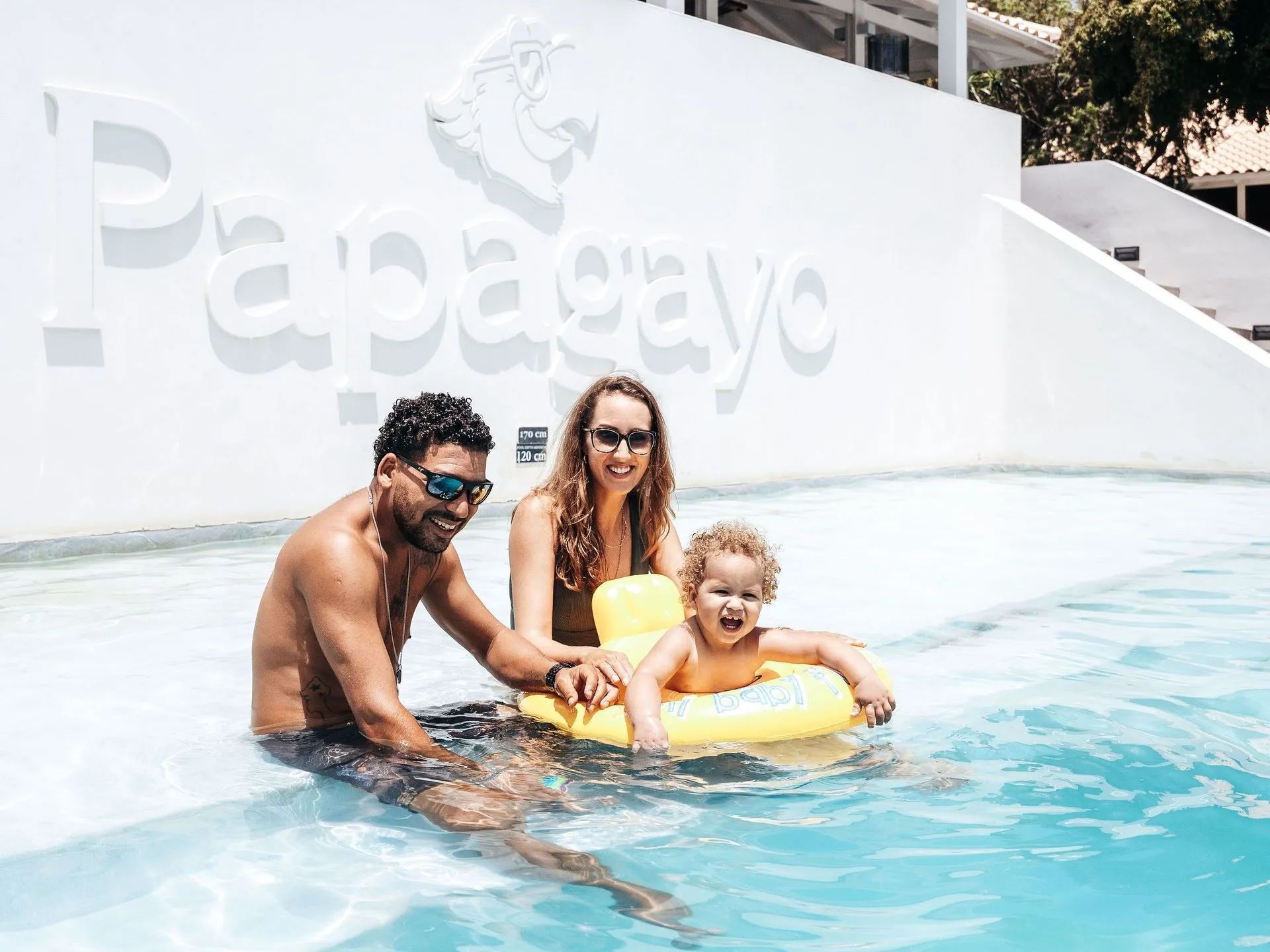 Papagayo Resort Pool Smiling family with a toddler enjoying the pool at Papagayo Beach Hotel & Resort in Curaçao, with the Papagayo logo in the background.