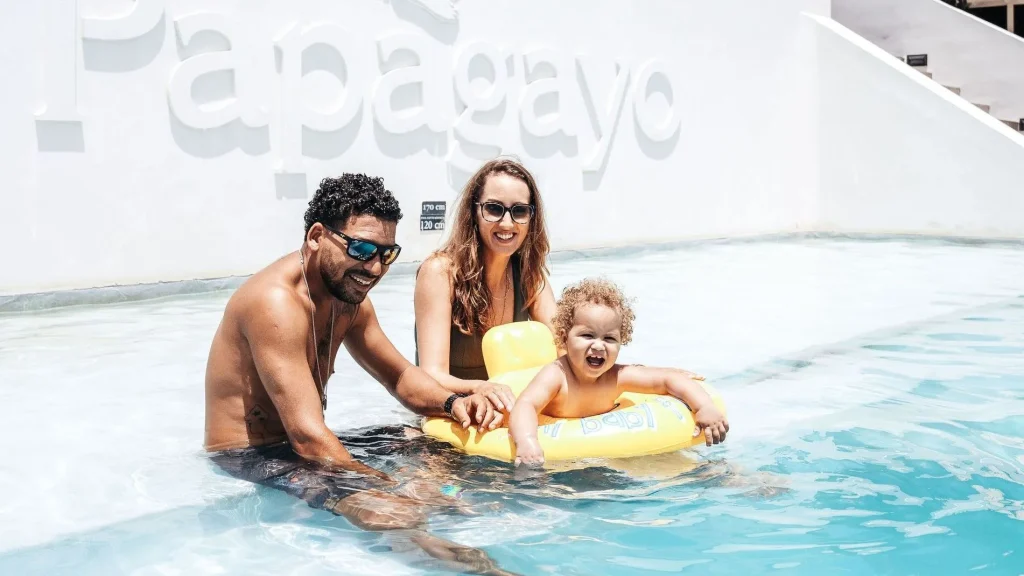 Smiling family with a toddler enjoying the pool at Papagayo Beach Hotel & Resort in Curaçao, with the Papagayo logo in the background.