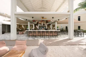 Open-air beach bar at Papagayo Curaçao with high stools, wooden deck flooring, soft lounge chairs on white sand, and tropical ceiling lanterns under a vaulted roof.