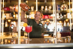 Smiling bartender at Papagayo Beach Hotel pouring a vibrant red cocktail into a glass, garnished with an orange slice, behind a stylish backlit bar.
