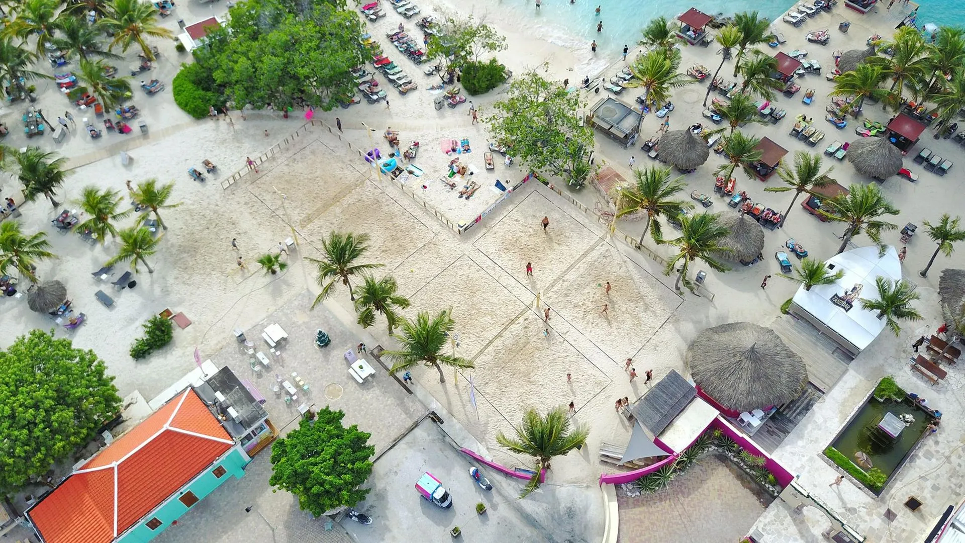 Aerial view of a vibrant beachside resort in Curaçao featuring multiple sandy beach volleyball courts surrounded by palm trees, sun loungers, and palapas, with guests relaxing by the turquoise shoreline.