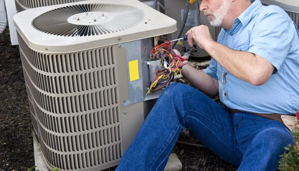 Papagayo AC technician repairing an outdoor air conditioning unit, using a screwdriver to work on exposed wiring while seated on the ground, dressed in a blue shirt, jeans, and a yellow hard hat.