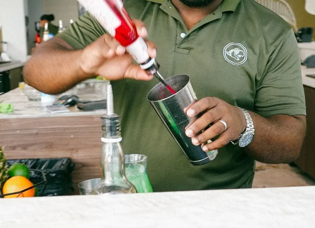 Papagayo bartender in a green uniform shirt pouring red syrup into a cocktail shaker at an outdoor bar, surrounded by fresh ingredients and bar tools.