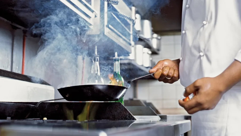 Close-up of a chef at Papagayo cooking in a professional kitchen, holding a frying pan over a gas flame with visible smoke rising, dressed in a white chef’s uniform with stainless steel kitchen equipment in the background.