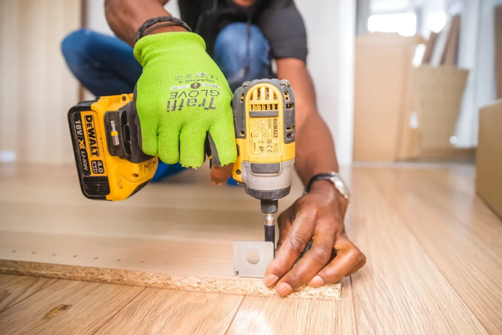 Close-up of a Papagayo handyman using a DEWALT power drill while assembling furniture on a wooden floor, wearing bright green safety gloves