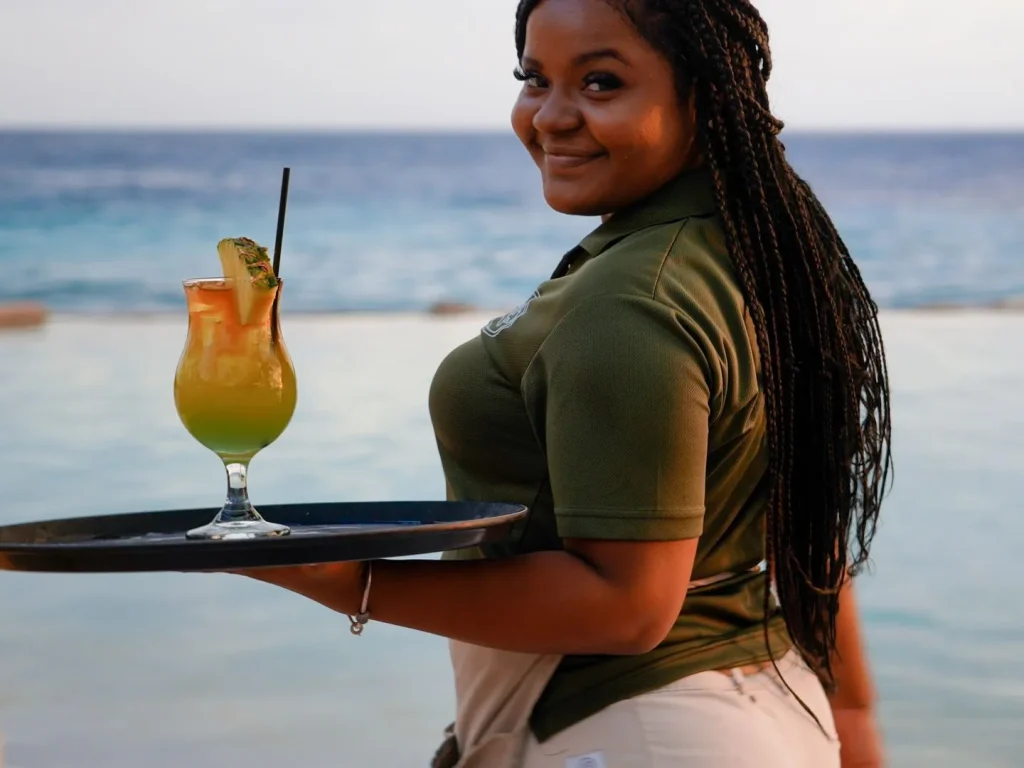Friendly Papagayo server with long braids smiling while holding a tray with a vibrant tropical cocktail garnished with pineapple, standing near the oceanfront pool at sunset