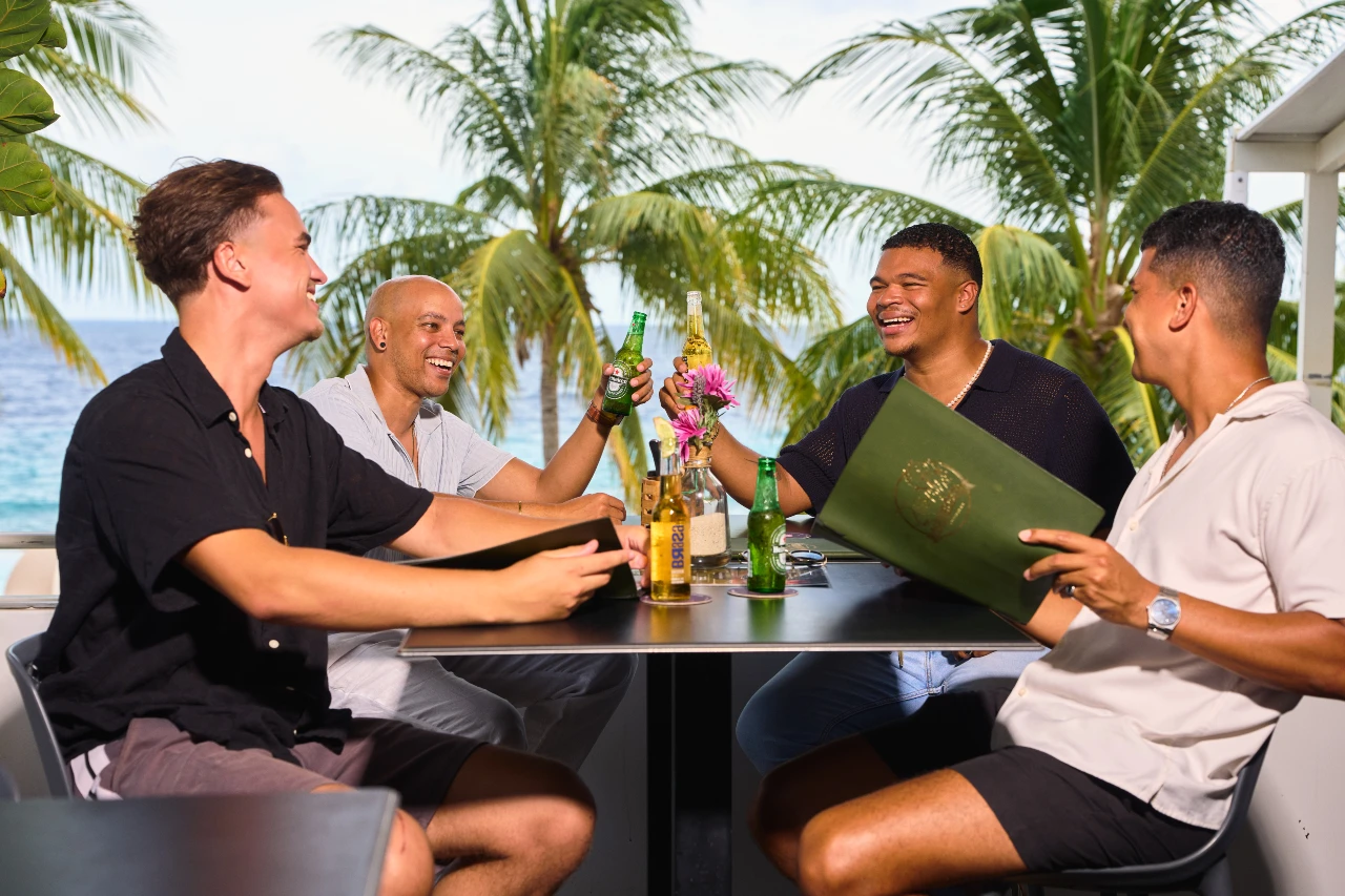 Four men share laughter and toast with beers on a sunny terrace at Papagayo Plaza at Mojitos, with tropical palm trees and the ocean in the background.