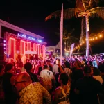 A vibrant crowd enjoys a nighttime concert at Papagayo Beach Club, with a brightly lit stage, palm trees, and string lights creating an electric atmosphere.
