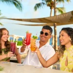 Three friends toast with vibrant cocktails under palm trees by the ocean at Papagayo Beach Club.