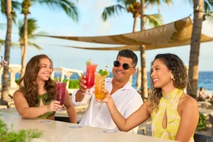 Three friends toast with vibrant cocktails under palm trees by the ocean at Papagayo Beach Club.
