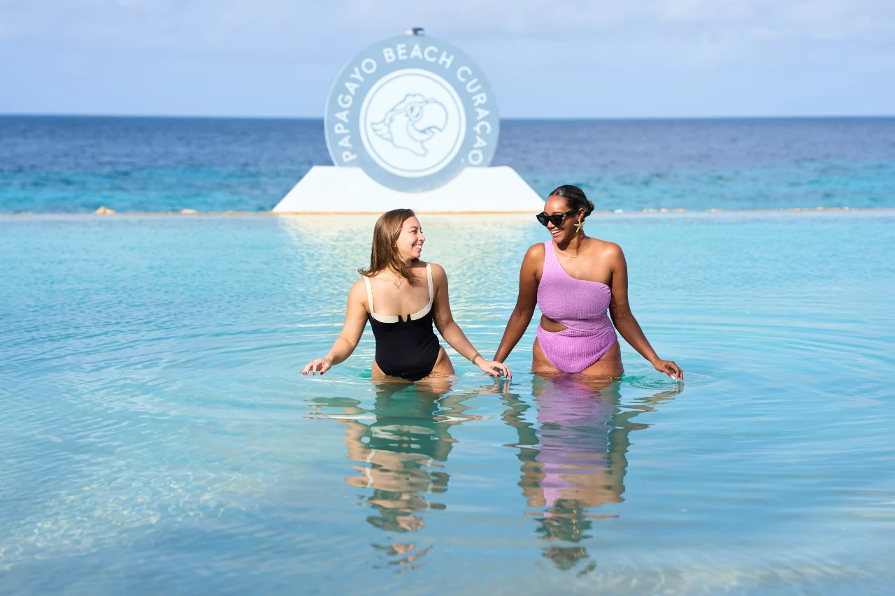 Two women relax in the infinity pool overlooking the Caribbean Sea, with the Papagayo Beach Curaçao logo in the background.