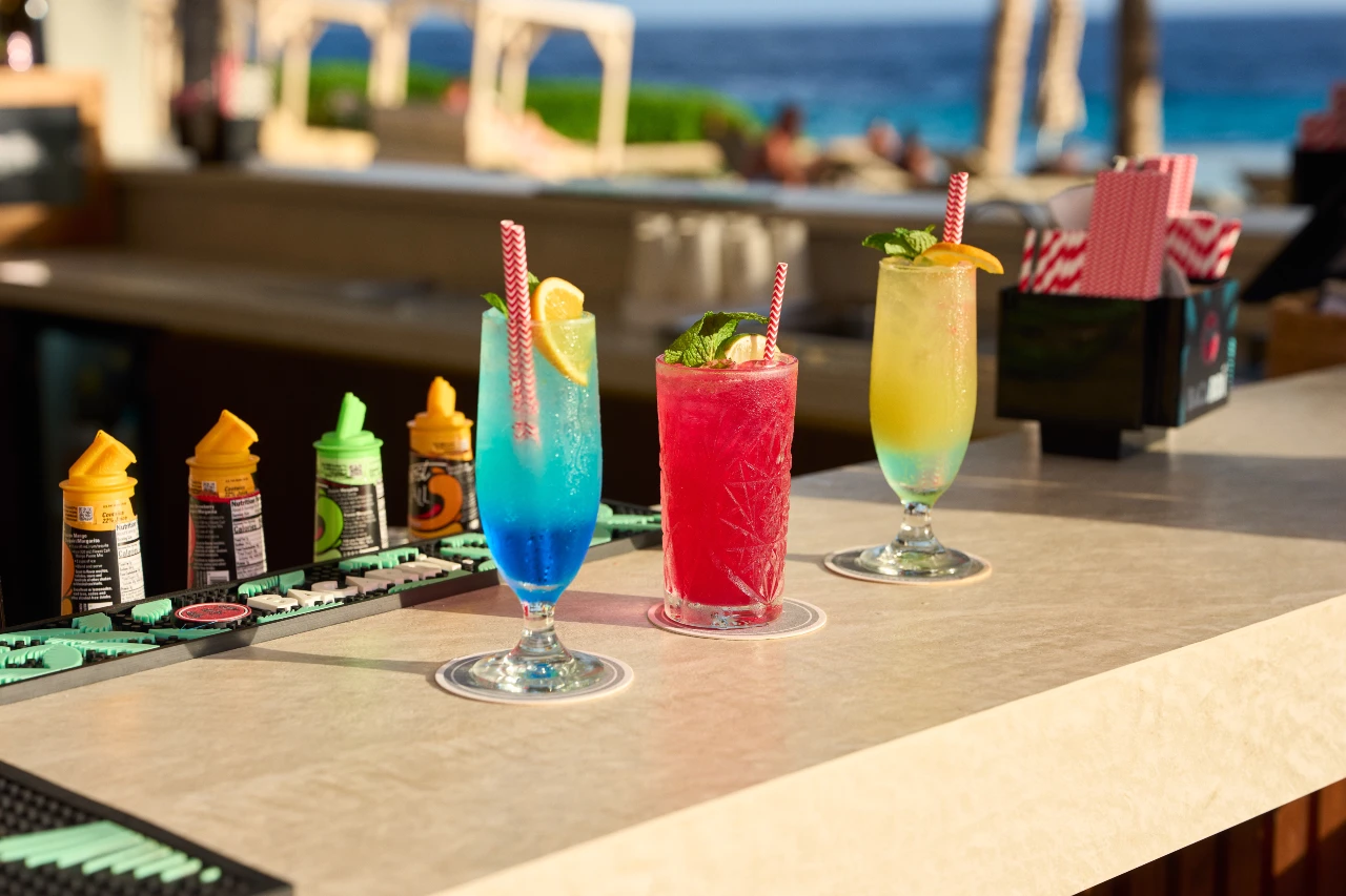 Papagayo Beach Club-ervaringen Drie kleurrijke cocktails op de bar, met de Caribische Zee op de achtergrond.