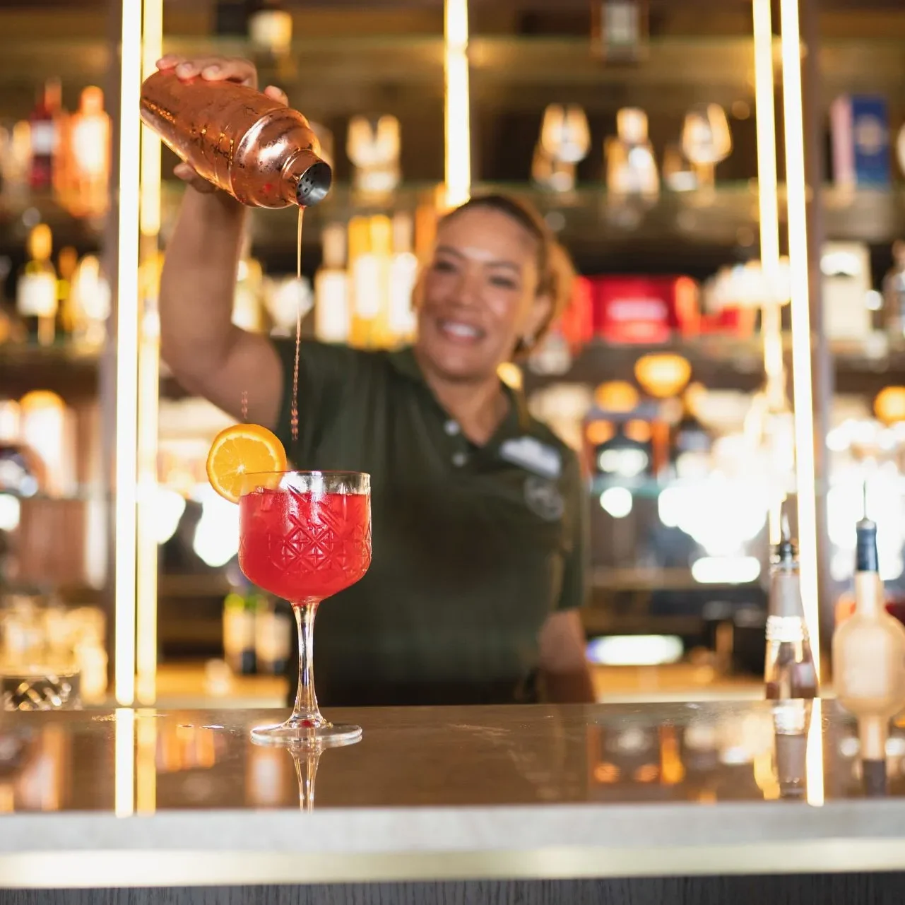 Papagayo Hotel Indoor Bar Smiling bartender at Papagayo Beach Hotel pouring a vibrant red cocktail into a glass, garnished with an orange slice, behind a stylish backlit bar.