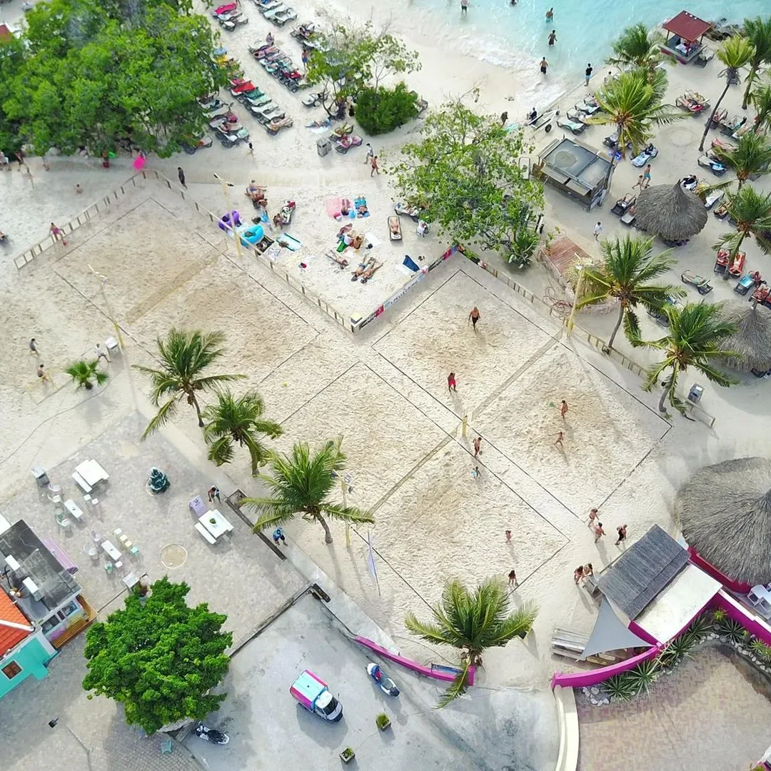Aerial view of a vibrant beachside resort in Curaçao featuring multiple sandy beach volleyball courts surrounded by palm trees, sun loungers, and palapas, with guests relaxing by the turquoise shoreline.